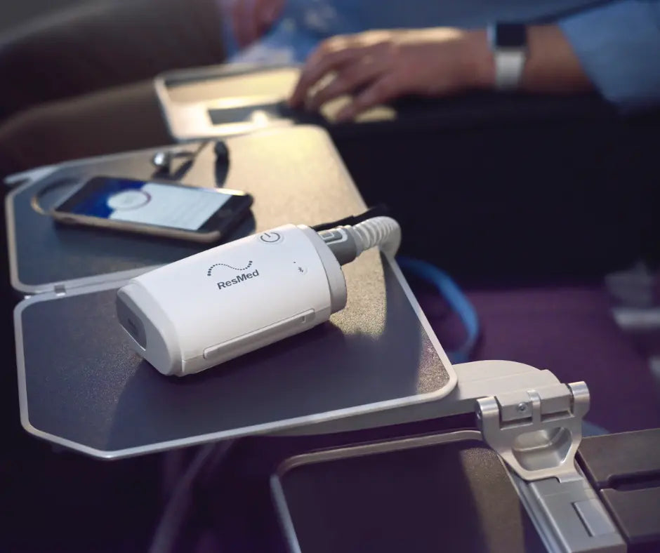 ResMed AirMini CPAP device set up on an airplane tray table beside a phone.