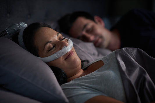 a woman comfortably sleeping while wearing AirFit P30i Nasal Pillow CPAP Mask, a man sleeping soundly in the background