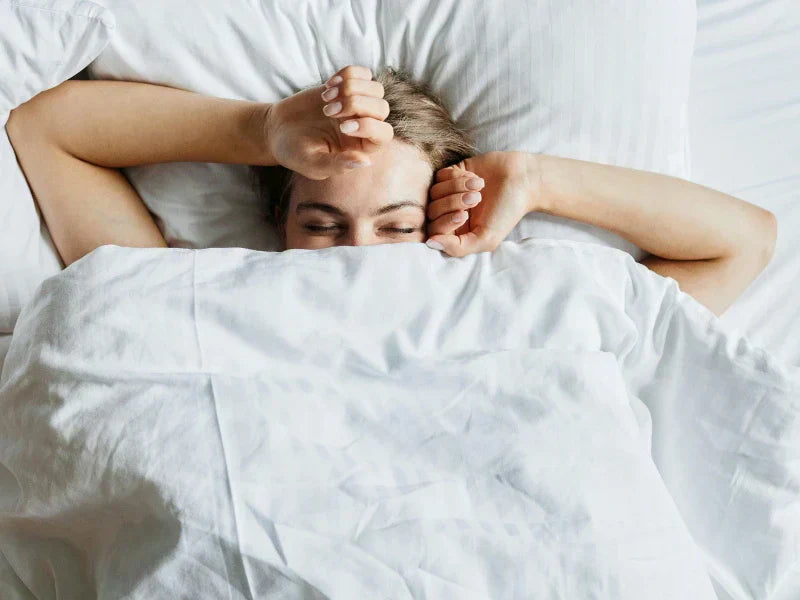  A person with closed eyes and hands over their face is lying in a bed with white sheets.